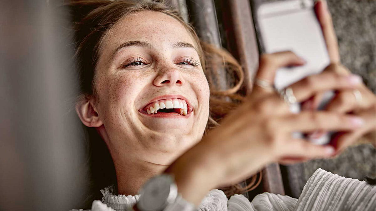 Eine junge Frau schaut lachend auf ihr Smartphone, dabei sieht man ihre schönen Zähne.